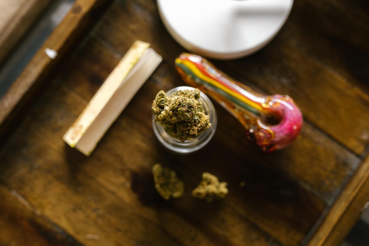 Top view of cannabis buds, a glass pipe, and rolling papers on a wooden surface.